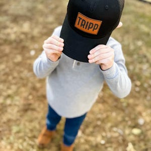 May include: A black mesh trucker hat with a brown leather patch that says "TRIPP" on it.