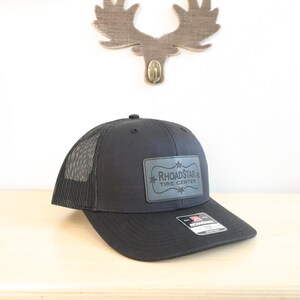 May include: A charcoal gray trucker hat with a mesh back and a curved brim. The hat features a rectangular patch with the text "Rhoadstar Tire Center" in a stylized font. A small tag is attached to the brim.