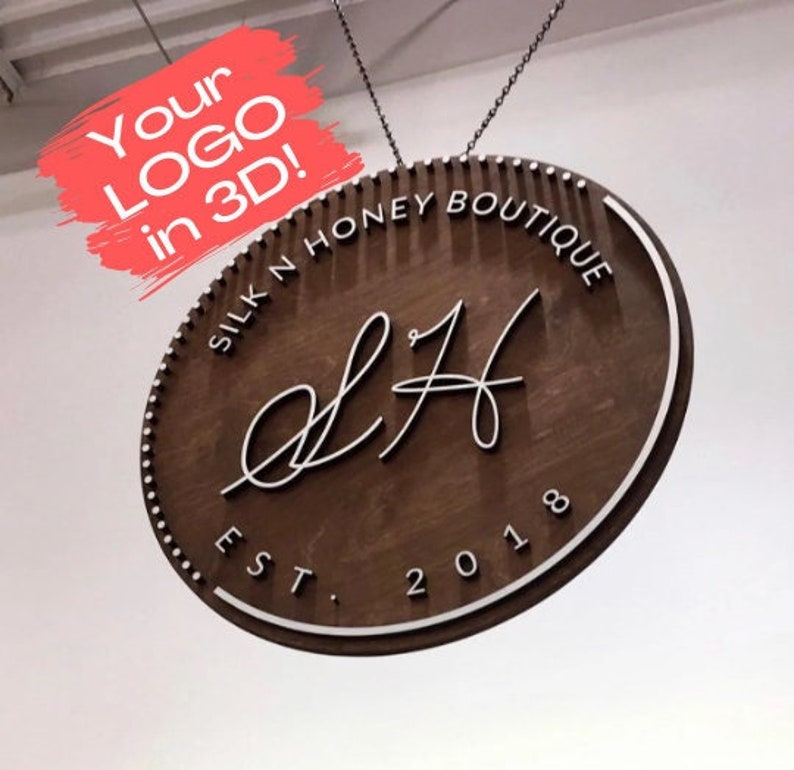 Op de afbeelding: Een bruin houten bord met een witte ovale rand en de tekst "SILK N HONEY BOUTIQUE" en "EST. 2018". Het bord heeft ook een wit cursief logo "L H".