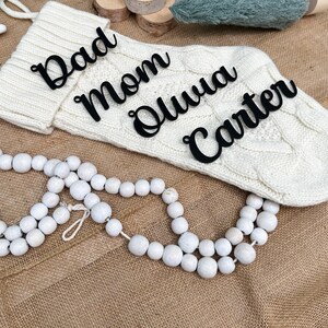 May include: White knitted stocking with black script lettering that reads "Dad", "Mom", "Olivia", and "Carter". The stocking is on a brown burlap surface with white wooden beads.