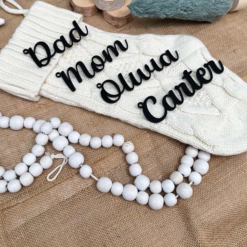 May include: White knitted stocking with black script lettering that reads "Dad", "Mom", "Olivia", and "Carter". The stocking is on a brown burlap surface with white wooden beads.