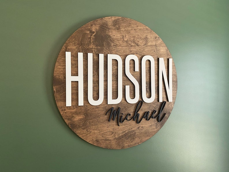 Op de afbeelding: Een rond houten bord met de naam "HUDSON Michael" in witte en zwarte letters.