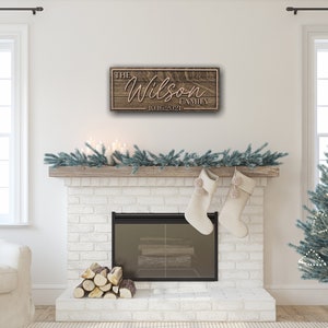 May include: A wooden sign with the text "The Wilson Family - 10.16.2021" hanging above a white brick fireplace with a garland of pine branches and white candles. There is a Christmas tree in the background.