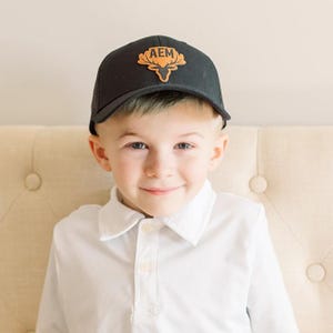 May include: A young boy wearing a black and orange baseball cap with a deer head logo and the letters "AEM" on it. He is wearing a white collared shirt.