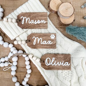 May include: Three wooden gift tags with white lettering. The tags are labeled "Mason", "Max", and "Olivia". The tags are on a white knitted surface with a string of white beads.