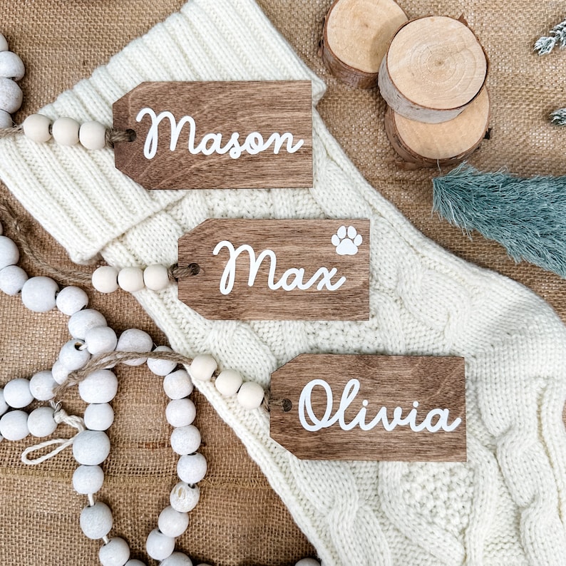 May include: Three wooden gift tags with white lettering. The tags are labeled "Mason", "Max", and "Olivia". The tags are on a white knitted surface with a string of white beads.