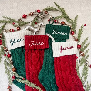 May include: Four red and green knitted Christmas stockings with white cuffs. The stockings are decorated with the names "Julian", "Jesse", "Lauren", and "Jaxson" in green embroidery.