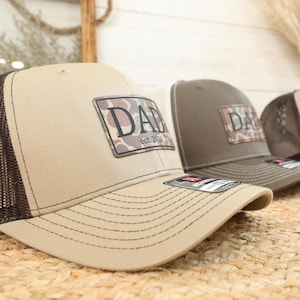 May include: Tan and brown trucker hats with a camouflage patch that reads "DAD" and "est. 2026". The hats have a mesh back and a curved brim. The hats are displayed on a woven surface.