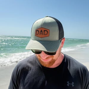 May include: A green and black trucker hat with the word "DAD" in a brown hexagon patch. The hat has a curved brim and a mesh back. The person in the image is wearing sunglasses and a black shirt.