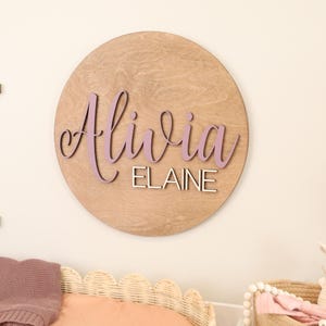 May include: A round wooden sign with the name "Alivia" in lavender script and the name "Elaine" in a smaller, block font. The sign is mounted on a wall, with a bed and basket visible below. The wood has a light brown color.