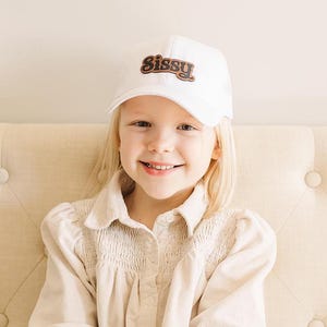 May include: A young person wearing a white baseball cap with the word "Sissy" in brown letters on the front. The person is wearing a light beige button-up shirt.