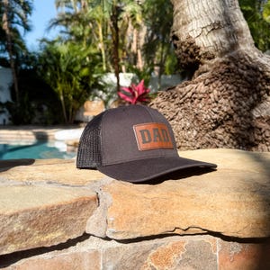 Puede incluir: Gorra de camionero negra con parte trasera de malla y visera curva. La parte delantera presenta un parche de cuero marr&oacute;n con la palabra "DAD". La gorra est&aacute; sobre una superficie de piedra.