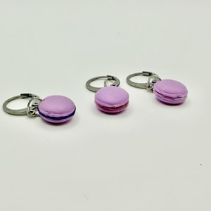 May include: Three stitch markers, each featuring a small, light purple macaron charm attached to a silver-toned metal ring. The macarons have a smooth, rounded top and bottom with a visible dark purple filling layer between the two halves.
