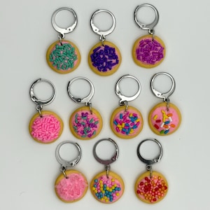 Pode incluir: Um conjunto de dez marcadores de pontos circulares, cada um com uma base de biscoito bege coberta com glacê rosa e granulados coloridos. Cada pingente de biscoito está preso a um aro de metal prateado, projetado para projetos de tricô ou crochê.