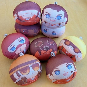May include: A collection of round plush keychains featuring various character designs. Each keychain has a different face and outfit, with a small metal chain for attachment. The keychains are primarily red, brown, orange, and yellow.