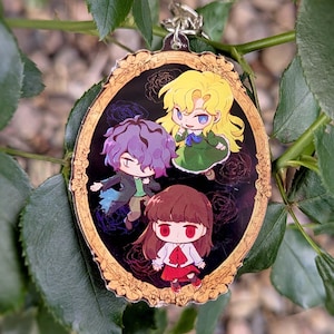 May include: An oval acrylic keyring featuring a gold-coloured ornate frame design. Inside the frame are three chibi-style characters with purple, blonde, and brown hair against a black background with subtle rose outlines and a silver metal ring.