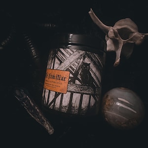 May include: A black candle jar with a white and black label that says "The Familiar" and "Vanilla Autumn Spice". The label also features a black cat illustration. The candle jar is sitting on a dark surface with a white bone and a gray orb.