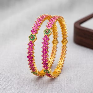 May include: Two gold bangles with a repeating pattern of pink gemstones. Green floral accents are interspersed along the bands. The bangles are displayed on a light gray surface.
