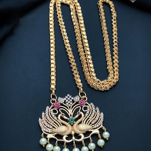 May include: A gold-toned necklace featuring a long, textured chain and a large pendant shaped like two facing swans. The pendant is embellished with clear white stones, pink accents, a central green stone, and seven dangling white pearls at the base.