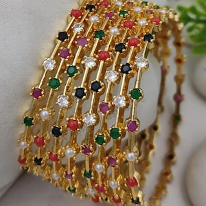 May include: Gold-toned bangles adorned with colorful gemstones and pearls. The bangles feature a repeating pattern of red, green, black, white, and clear stones. The jewelry is displayed on a white surface.