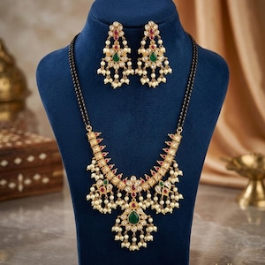 May include: A gold-toned necklace and earring set featuring black beads, white pearls, red stones, and green teardrop-shaped accents. The necklace has a central pendant with dangling pearls, and the matching earrings mirror this intricate design.