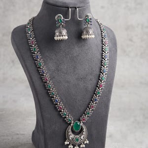May include: A long silver-toned necklace featuring a central crescent-shaped pendant with a green oval stone, accented by smaller green, blue, and pink teardrop stones. Matching jhumka-style earrings with green stones and white bead dangles.