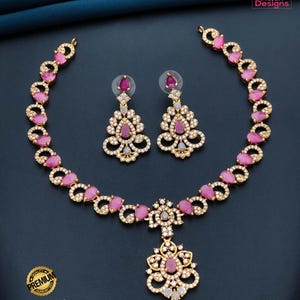 May include: A gold-toned necklace and earring set with pink gemstones and clear crystals. The necklace features a floral pendant and matching earrings. The set is displayed on a dark blue background. The text "Premium Quality Matte Finish AD Necklace" is visible.