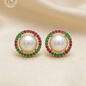 May include: A pair of round stud earrings featuring a central white pearl surrounded by a circular halo of alternating small green and pink gemstones set in gold. The earrings are displayed with a logo in the corner reading Indian Designs.