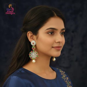 May include: Elegant dangle earrings featuring a circular top with white stones and a red center stone. The earrings have a floral design with red, green, and white stones, and a large pearl at the bottom. The earrings are gold-toned and are worn by a person.