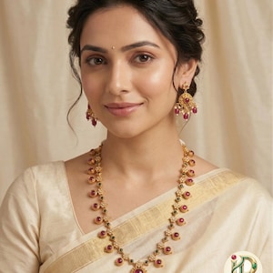 May include: A woman wearing a gold-toned necklace and matching earrings. The jewelry features red and green gemstones and small white pearls. The woman is wearing a cream-colored garment with gold trim. The image is well-lit and the jewelry is the focal point.