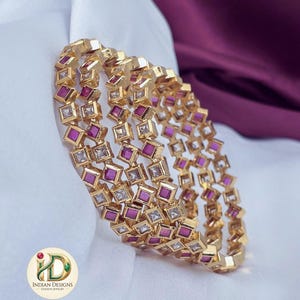May include: A set of three gold-toned bangles featuring a repeating square pattern. Each square is set with either a clear white stone or a vibrant magenta-pink stone. The jewelry includes a logo with the text Indian Designs Fashion Jewelry.