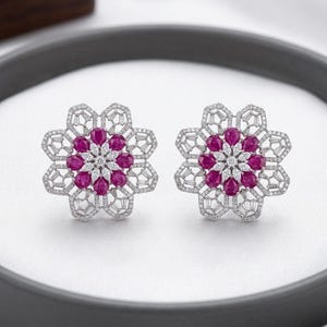 May include: A pair of flower-shaped stud earrings. Each earring features a central cluster of pink gemstones surrounded by white gemstones and a silver-colored, openwork frame. The earrings are displayed on a white surface.