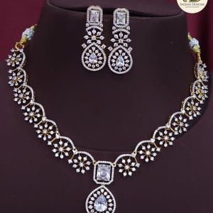 May include: A gold-coloured necklace and earring set. The necklace has a central square-cut stone with a teardrop pendant, and is decorated with white gemstones. The matching earrings have a similar design, with rectangular and teardrop-shaped stones.
