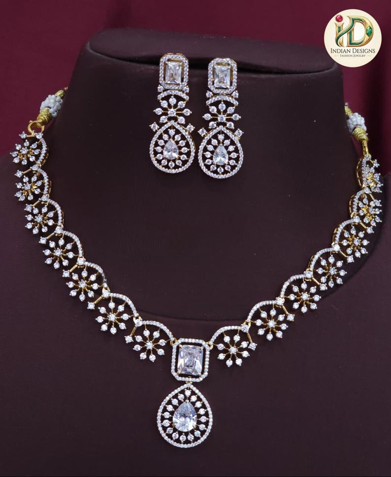 May include: A gold-coloured necklace and earring set. The necklace has a central square-cut stone with a teardrop pendant, and is decorated with white gemstones. The matching earrings have a similar design, with rectangular and teardrop-shaped stones.