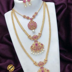 May include: A gold-toned jewelry set featuring a multi-layered necklace and matching earrings. The pieces are adorned with small pink stones and white pearl-like beads. The necklace has three distinct chains with ornate, stone-encrusted pendants.