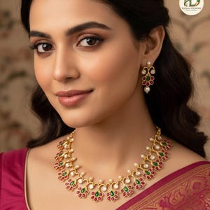 May include: Gold-toned necklace and matching earrings set. The jewelry features a floral design with red and green gemstones, accented by small white pearls. The necklace is worn on a red garment.