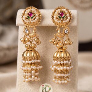May include: Gold-toned earrings with a floral design and a central red stone. The earrings feature a peacock motif, dangling gold beads, and multiple strands of small, white pearls. The earrings are displayed on a cream-colored surface.
