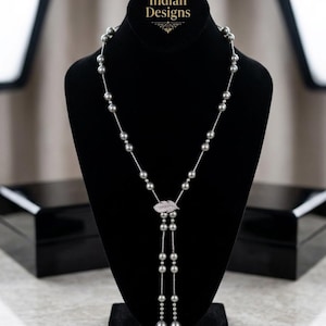 May include: A long necklace featuring grey spherical beads strung on a thin silver-toned chain. The necklace includes a central silver-toned leaf-shaped pendant encrusted with small clear stones. The text Indian Designs is visible at the top.