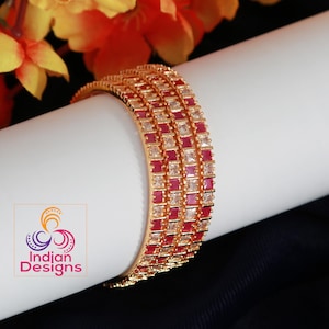May include: Set of four gold bangles with red and clear square-shaped stones. The bangles are stacked on top of each other.