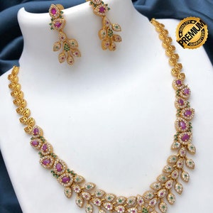 May include: A gold-toned necklace and earring set. The necklace and earrings feature a leaf and floral design with red, green, and clear gemstones. The earrings dangle from a small stud. The necklace is displayed on a white bust.