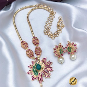 May include: A gold-tone necklace and earring set featuring a peacock-shaped pendant and matching earrings adorned with pink and green stones and white pearls. The necklace has a chain with a lobster clasp and the text Indian Designs and Premium Quality Micro Plated Necklace is visible.