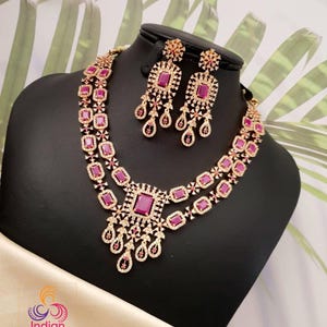 May include: A gold-toned necklace and earring set with ruby-colored rectangular stones and clear accent stones. The necklace has a layered design with a large central pendant. The earrings mirror the necklace's design. The set is displayed on a black mannequin.