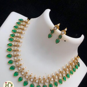 May include: A gold-toned necklace and matching earrings set. The necklace features teardrop-shaped green gemstones, small white pearls, and clear crystals. The earrings have a similar design with three green gemstones each. The jewelry is displayed on a white mannequin.