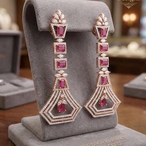 May include: Rose gold geometric drop earrings with ruby-colored stones and clear crystals. The earrings are displayed on a gray velvet stand. The text on the stand reads "GEOMETRIC DROP EARRINGS in 18K ROSE GOLD."