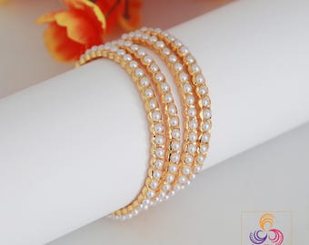 May include: Four gold bangles with white pearl accents are displayed on a white surface. The bangles are stacked together, showcasing their intricate design. The background features blurred orange and yellow flowers, and a logo for Indian Designs.