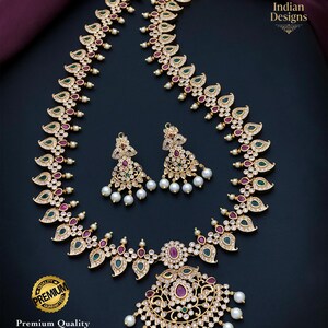 May include: A gold-toned necklace and earring set featuring a matte finish, adorned with teardrop-shaped green and red stones, clear crystals, and dangling white pearls. The necklace includes a large central pendant and the text Indian Designs.