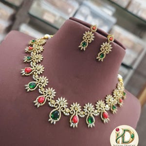 May include: A gold-toned necklace and earring set with red and green gemstones. The necklace and earrings feature a floral design with clear crystals. The jewelry is displayed on a brown mannequin head.
