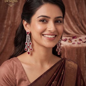 May include: A woman wearing long, dangling earrings with ruby-red rectangular stones and small, clear stones. The earrings are set in a gold-colored metal. The woman is wearing a brown top and a maroon sari. The text "Indian Designs" is in the upper left corner.