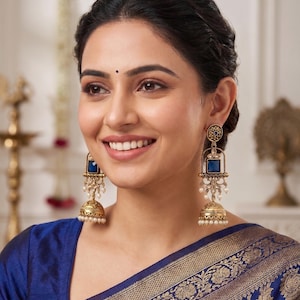 May include: A pair of gold-toned jhumka earrings featuring a square blue stone at the top, intricate metal filigree work, and a bell-shaped base adorned with small white pearl-like beads hanging from the bottom edge of the earring structure.