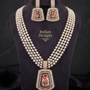May include: A pearl necklace and earring set. The necklace has multiple strands of white pearls and a rectangular pendant with a floral design. The earrings are square-shaped with a matching design. The text "Indian Designs" is visible.